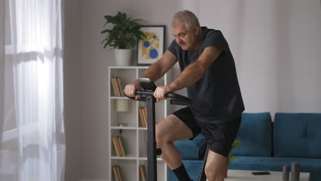 Müder-Mann-Trainiert-Zu-Hause-Mit-Einem-Stationären-Fahrrad.-Mittlere-Aufnahme-Einer-Grauhaarigen-Person-Mit-Schnurrbart,-Cardio-Training